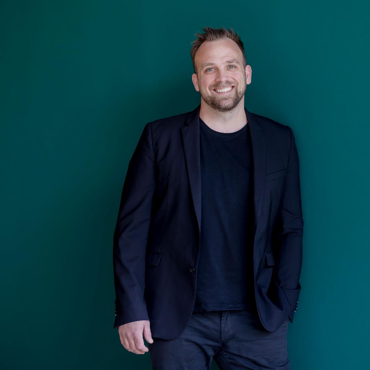 Portrait von DJ Dominik, der mit einer Hand in der Hosentasche vor einer dunkelgrünen Wand steht