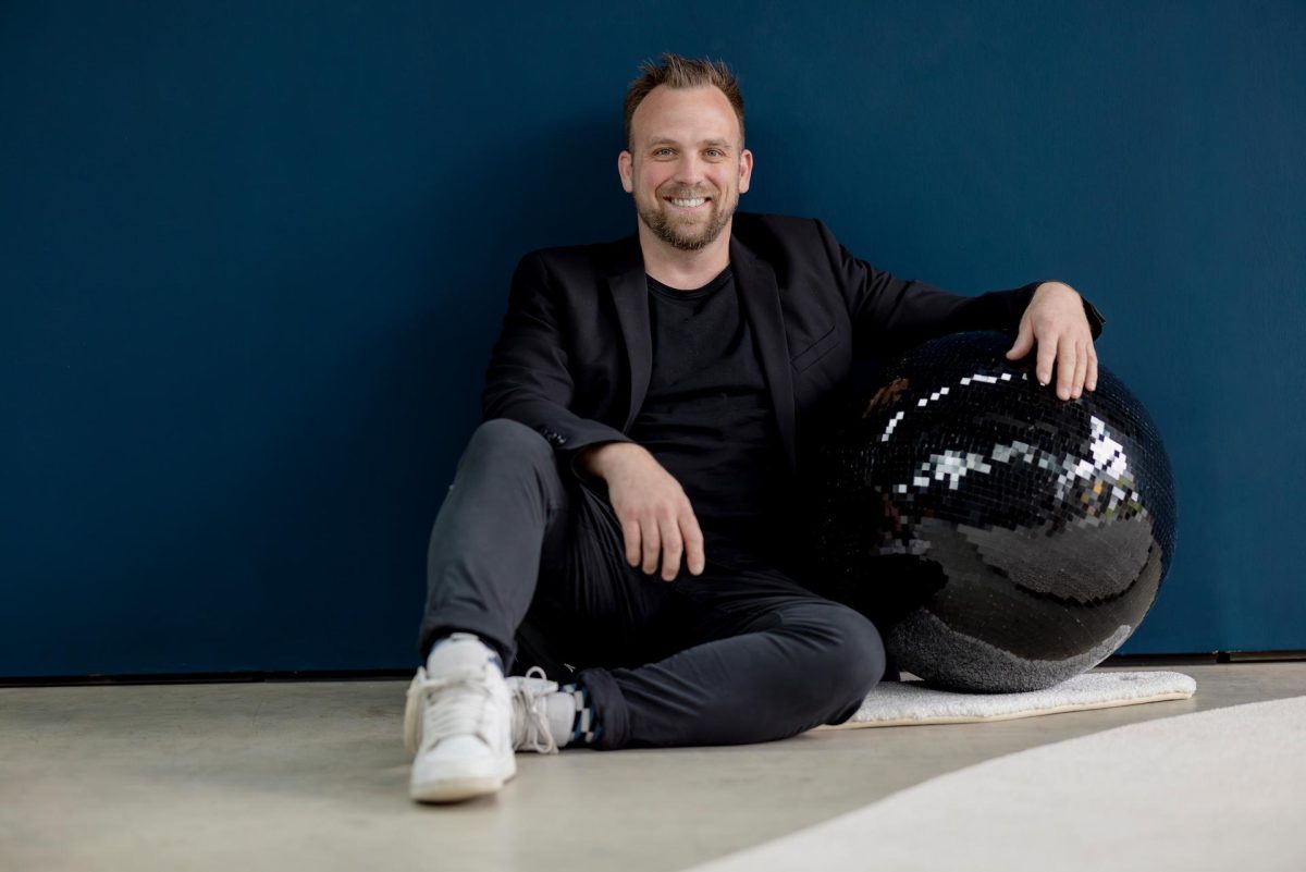 Portraitfoto von DJ Dominik, der mit einer schwarzen Disco Kugel unterm Arm vor einer blauen Wand sitzt