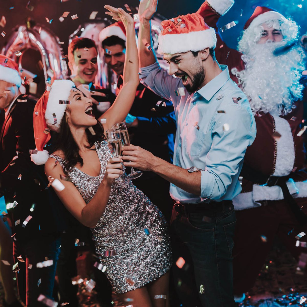 Guests at a Christmas party are wearing Santa hats and toasting with sparkling wine amidst a shower of confetti.
